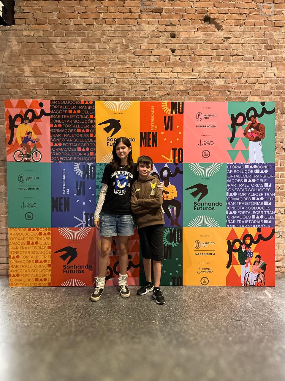 Maria Clara e Francisco diante do backdrop da décima edição do PAI na Pinacoteca